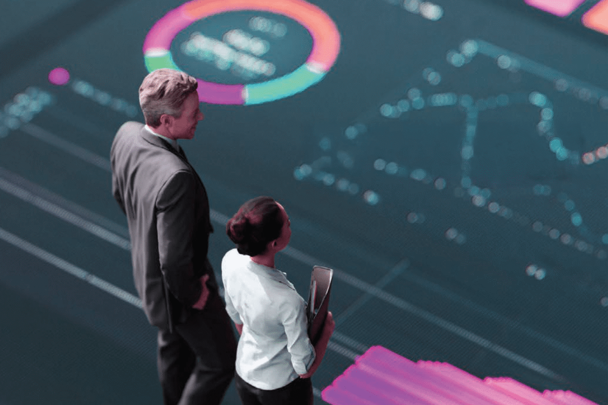Two business professionals standing together, reviewing a large, glowing digital dashboard displaying graphs and analytics.