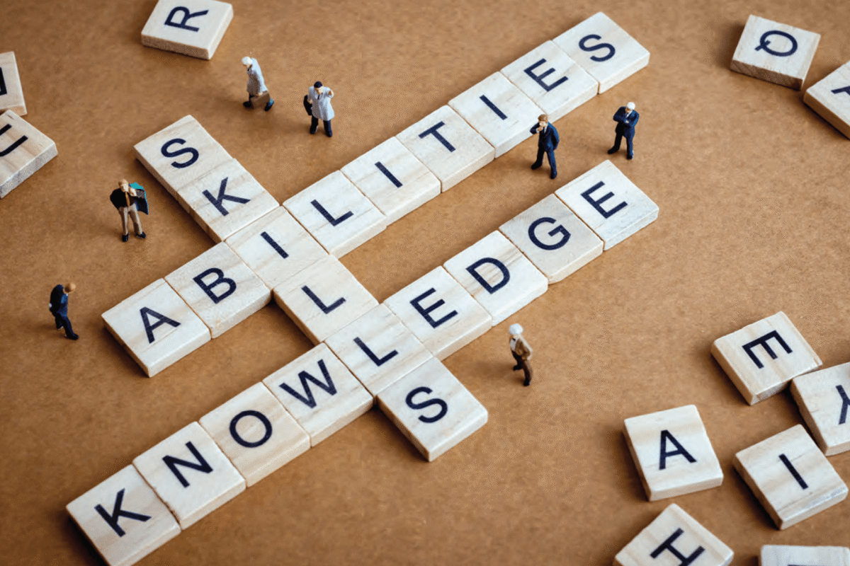 Letter tiles spell "abilities," "skills," and "knowledge" in a crossword-style arrangement, surrounded by miniature people, representing professional competencies and expertise.